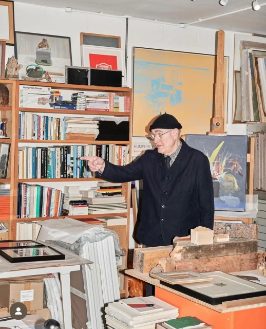 Elderly man in glasses and a black hat pointing in an artist studio with paintings, books, and various art supplies on shelves and tables.