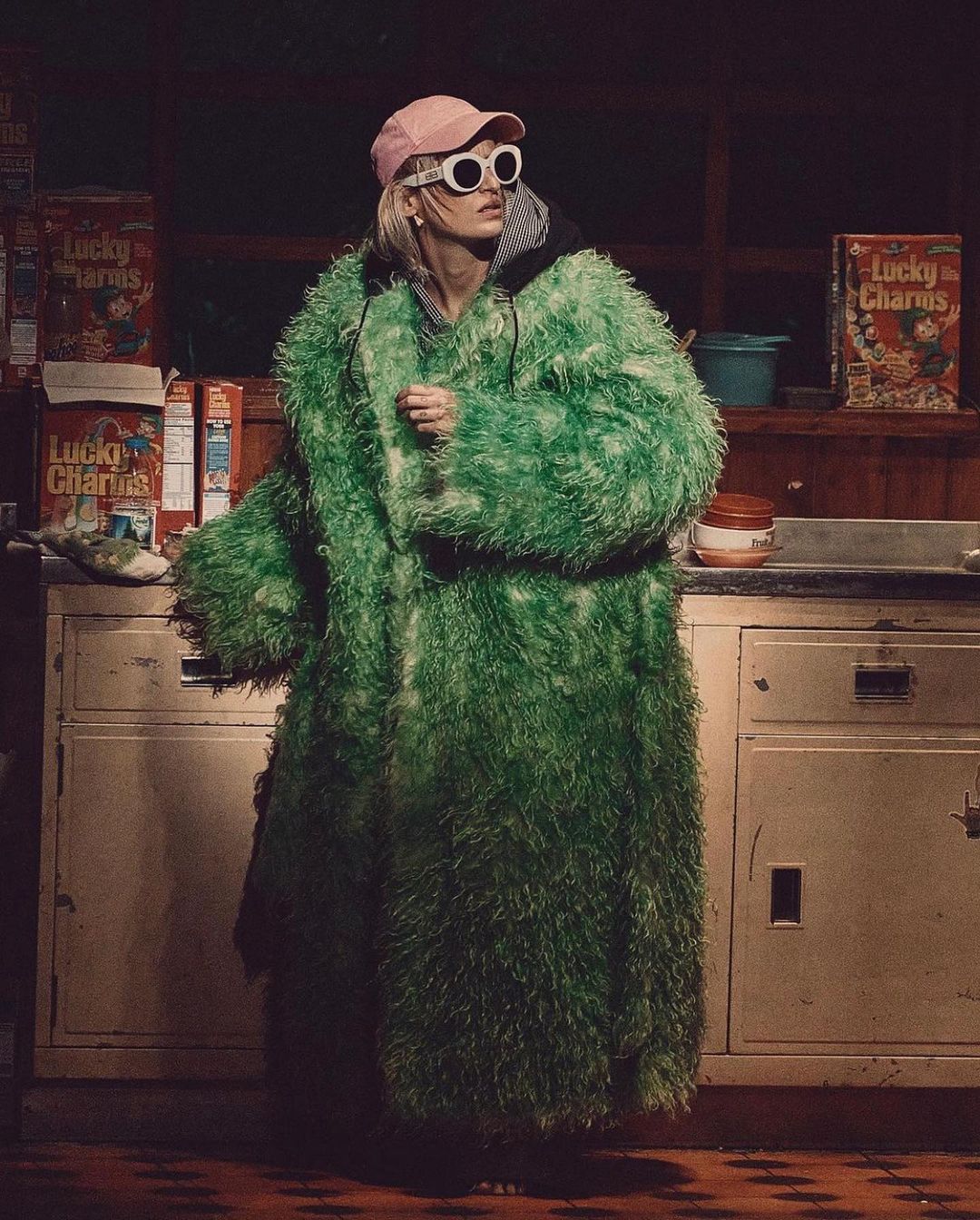 Adult woman in a large green faux fur coat, pink cap, and white sunglasses standing in front of Lucky Charms cereal boxes.
