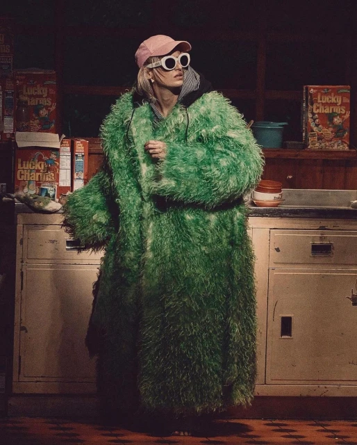Adult woman in a large green faux fur coat, pink cap, and white sunglasses standing in front of Lucky Charms cereal boxes.