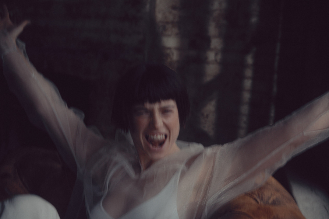 An excited adult female with short, dark hair wearing a white top and sheer jacket raises her arms in an indoor setting against a dark background.