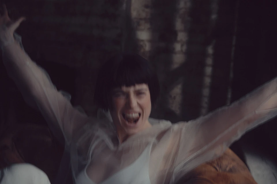 An excited adult female with short, dark hair wearing a white top and sheer jacket raises her arms in an indoor setting against a dark background.
