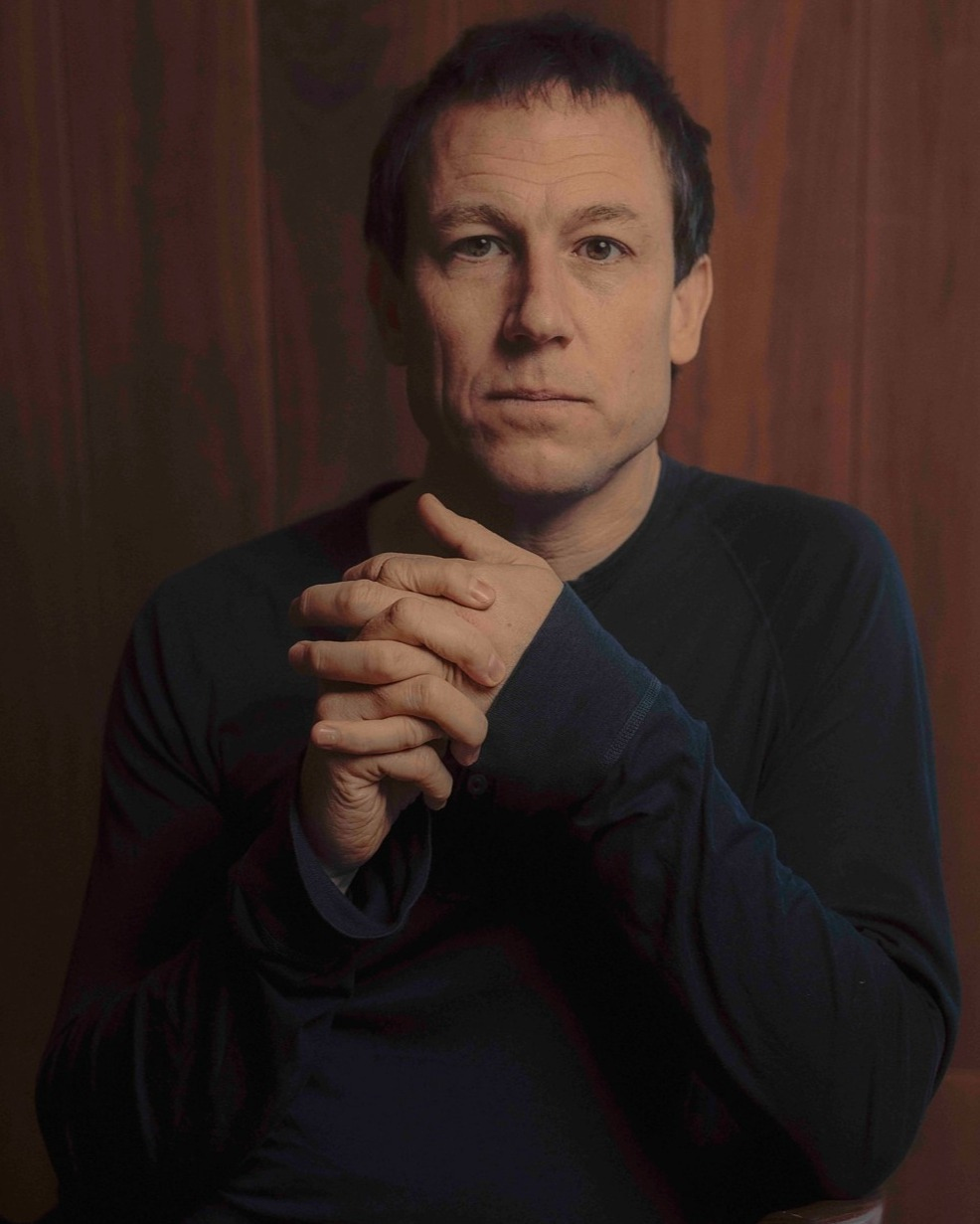 Portrait of a middle-aged man wearing a dark crewneck shirt with clasped hands against a dark wood paneling background.