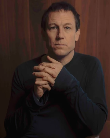 Portrait of a middle-aged man wearing a dark crewneck shirt with clasped hands against a dark wood paneling background.