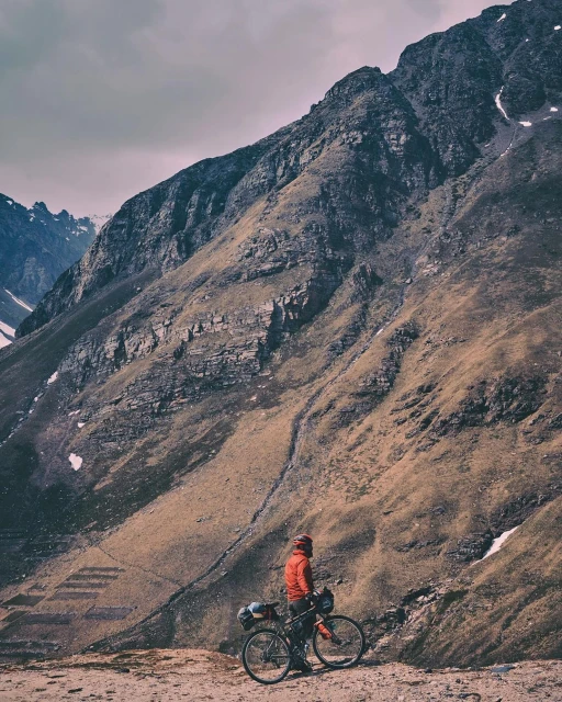 Adult male on a bicycle stands near a large mountain with sparse vegetation under a cloudy sky in an eye-level outdoor shot.