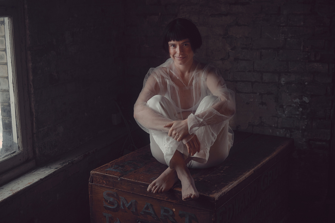 Young woman in sheer white top and pants sits cross-legged on a vintage trunk in dimly lit room with exposed brick and worn window.