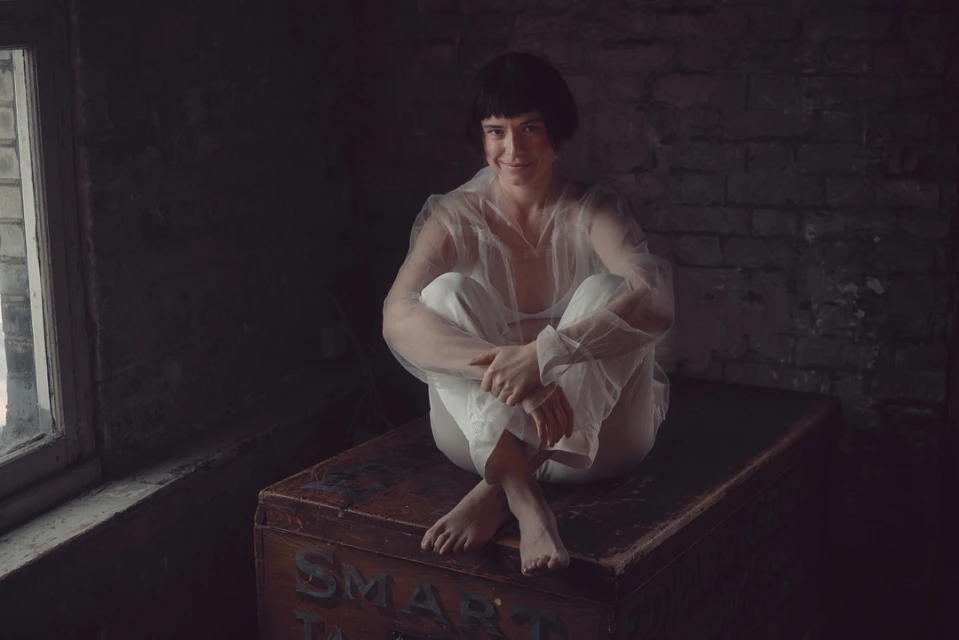 Young woman in sheer white top and pants sits cross-legged on a vintage trunk in dimly lit room with exposed brick and worn window.