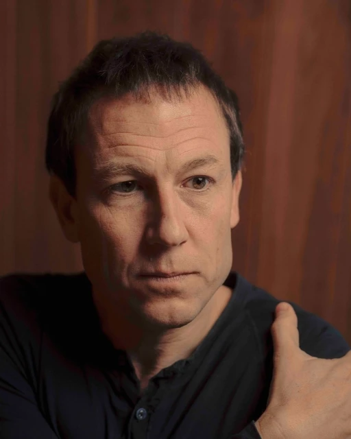 Close-up portrait of a mature adult male with short hair, wearing a black shirt, set against a neutral, dark brown backdrop in soft lighting.