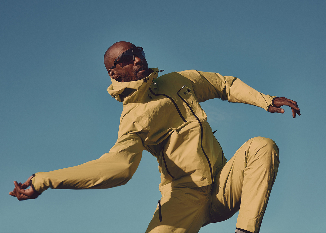 Black man in yellow athletic jacket and pants leaping against a clear blue sky