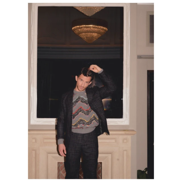 Man poses indoors with a hand in his hair wearing a dark suit with a patterned sweater in front of a white fireplace mantel.