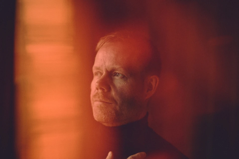Portrait of an adult male with short red hair, wearing a black turtleneck sweater against an orange and dark background.