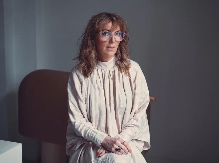 Portrait of an adult woman with wavy auburn hair wearing glasses and a beige dress, seated indoors against a neutral background.