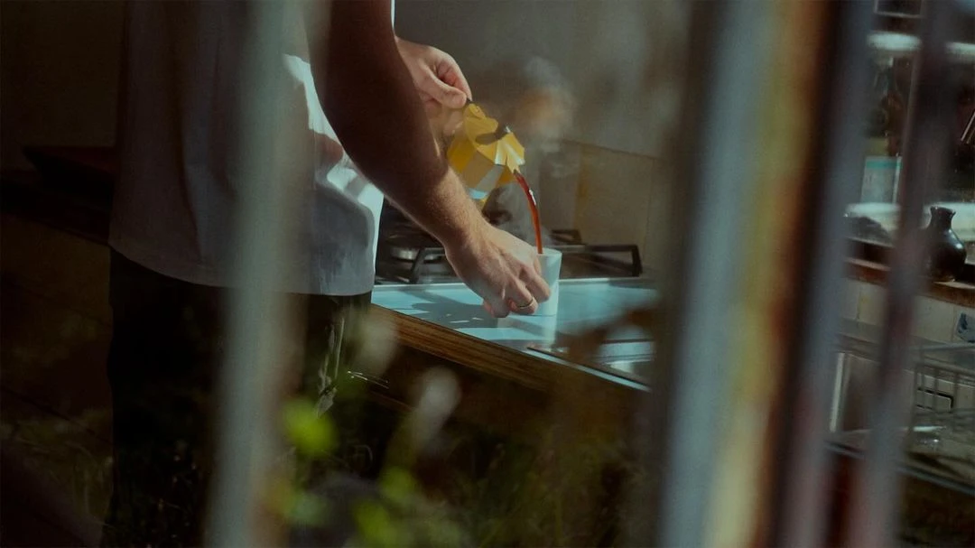 Man pours dark liquid from a yellow Moka pot into a white mug in an indoor kitchen, light streams in from a window.