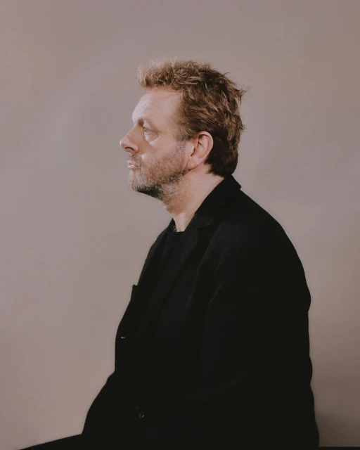 Side profile portrait of a middle-aged man with curly hair wearing a black jacket against a neutral background in a photo studio.
