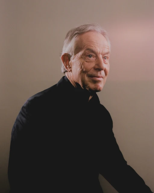 Portrait of an elderly white male with gray hair wearing a black collared shirt, looking off to the side against a neutral background.