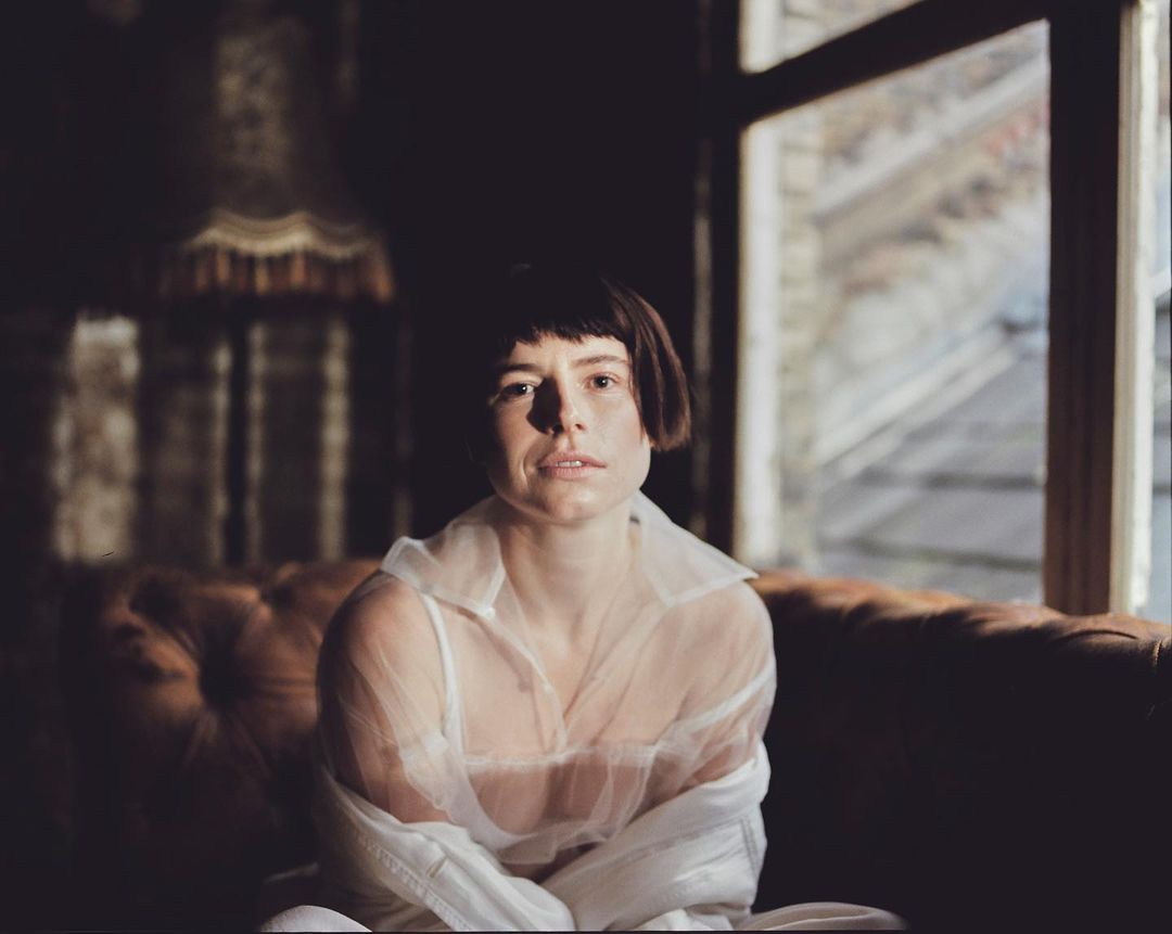 Indoor portrait of a young adult woman with short dark hair in a sheer white shirt, seated on a brown couch near a window.