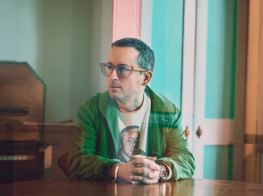 Portrait of a seated man with eyeglasses, wearing a green jacket and printed t-shirt, with hands clasped at a wooden table indoors.