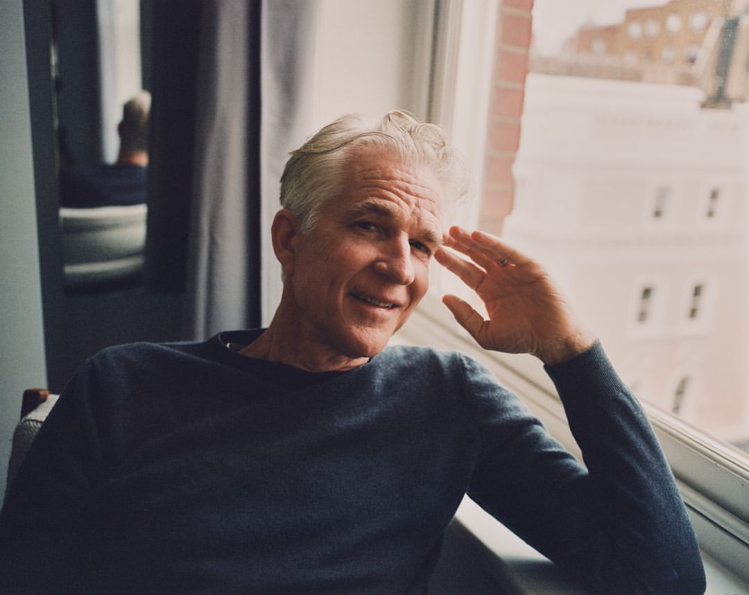 Portrait of a mature adult male with salt and pepper hair, wearing a blue sweater, sitting by a window with an urban background.