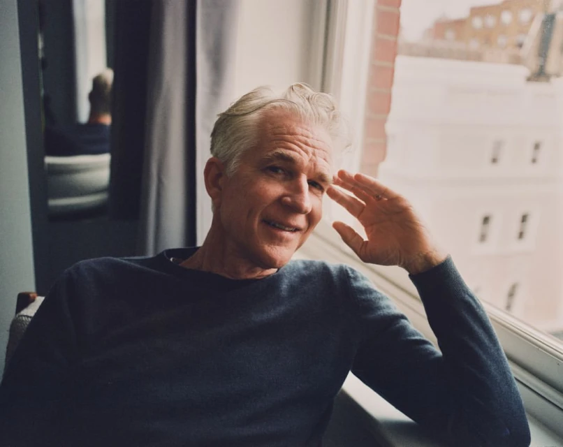 Portrait of a mature adult male with salt and pepper hair, wearing a blue sweater, sitting by a window with an urban background.