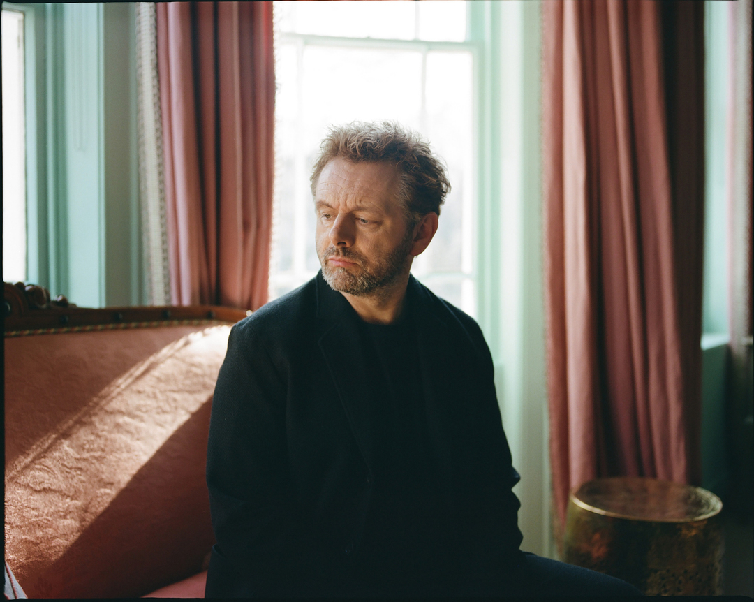 Portrait of a light-skinned adult male seated in a room with pink and light-green walls, and a large window letting in natural light.