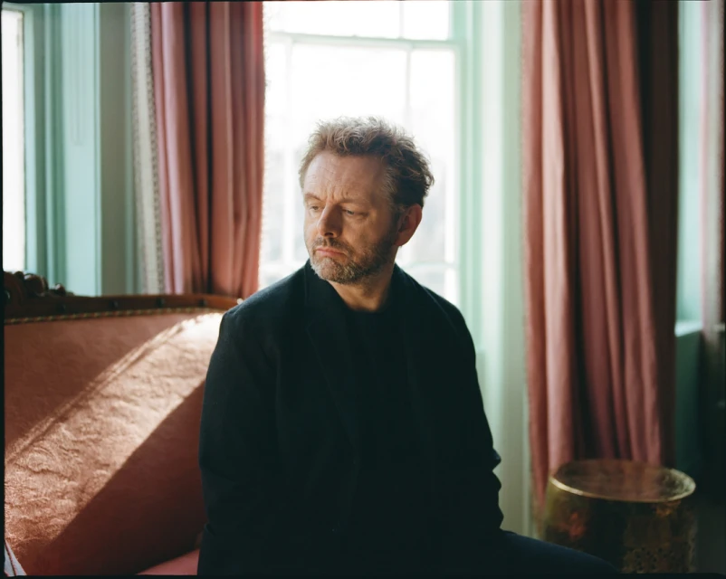 Portrait of a light-skinned adult male seated in a room with pink and light-green walls, and a large window letting in natural light.