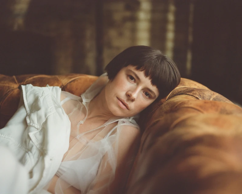 Portrait of a woman with short dark hair reclining on an amber-colored couch with a sheer white top and a white jacket, indoors.