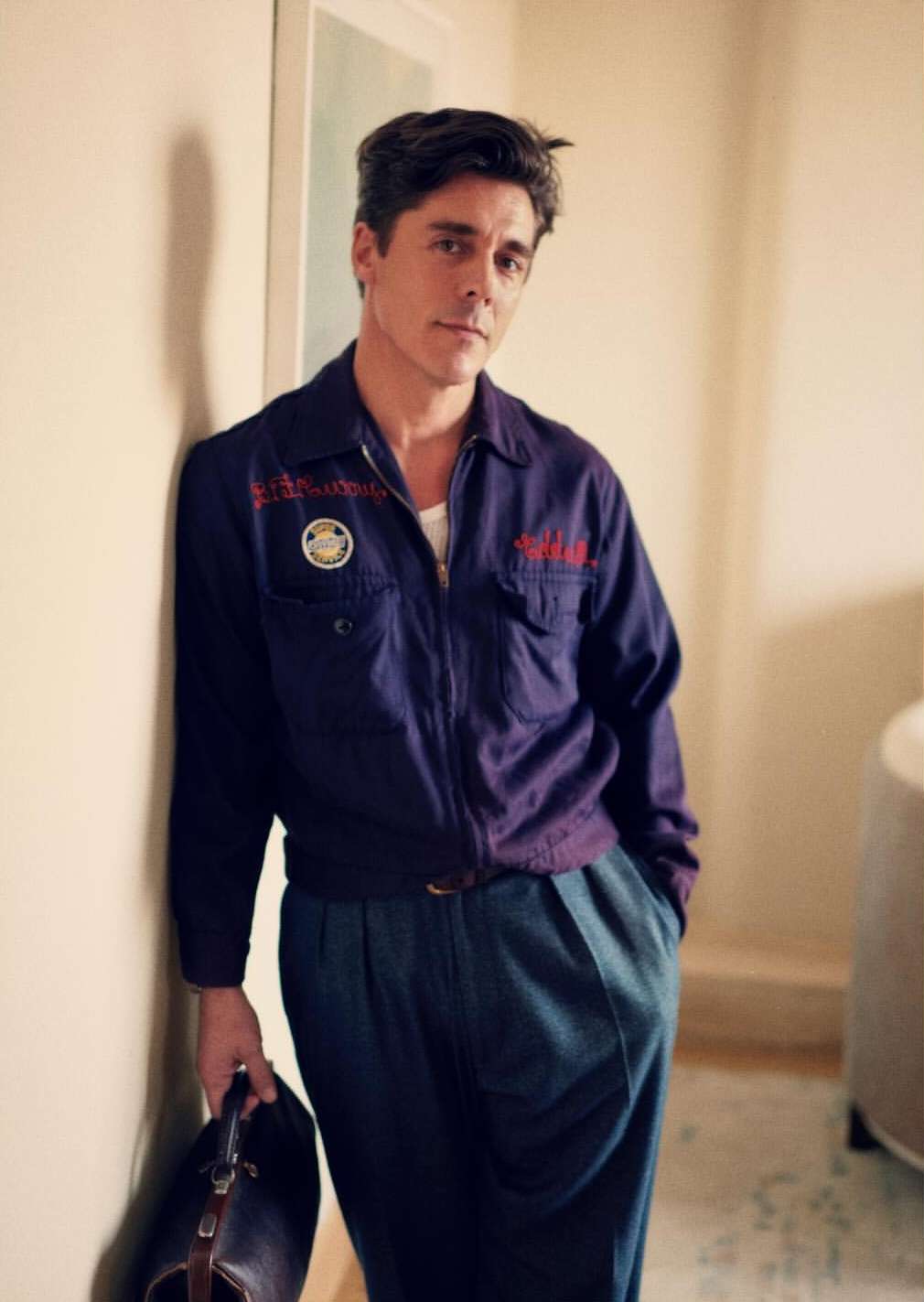 Portrait of a man in vintage workwear holding a bag, leaning against a wall indoors with soft, natural lighting.
