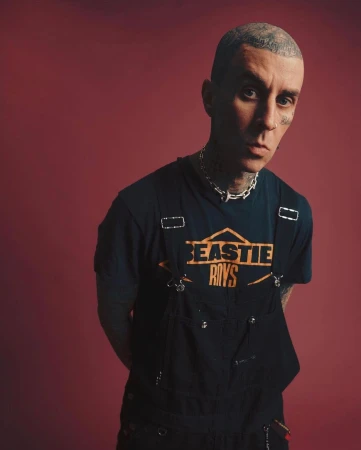 Studio shot of a tattooed adult male wearing a Beastie Boys t-shirt and black overalls against a red backdrop, looking at the camera.