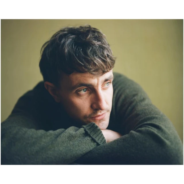 Portrait of a young adult male with curly dark hair, wearing a dark green sweater, resting his chin on his folded arms, indoors.