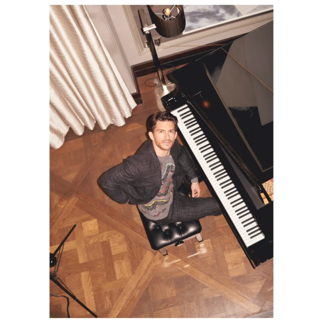 Man sits on piano bench in herringbone suit, view from above. Modern piano, floor lamp, and wood floor complete the composition.