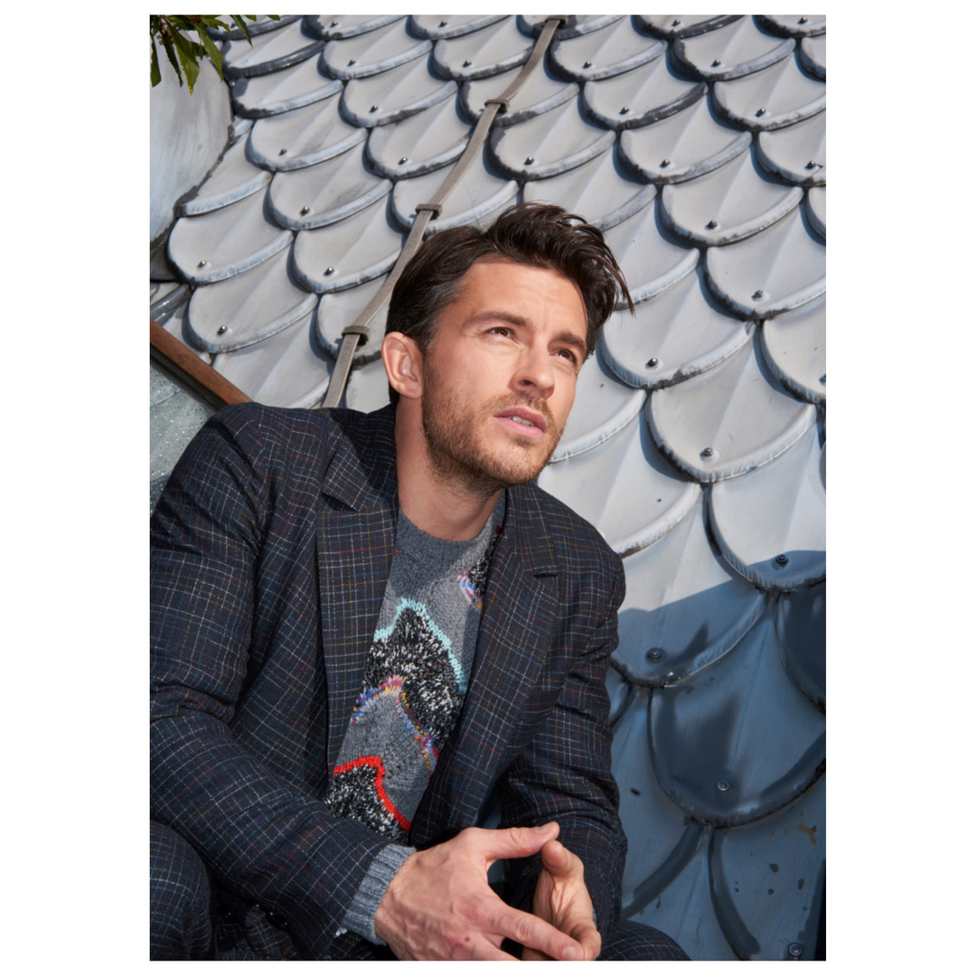 Portrait of a stylish adult male in a plaid blazer and patterned sweater, sitting outdoors against a textured tiled roof backdrop.