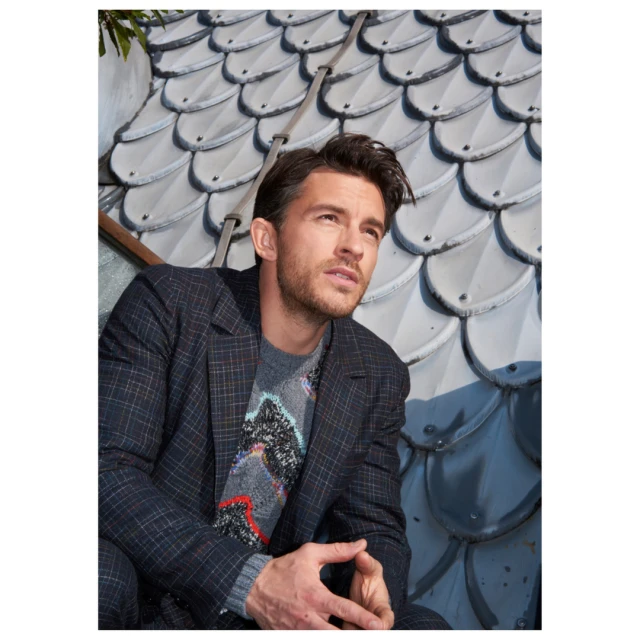Portrait of a stylish adult male in a plaid blazer and patterned sweater, sitting outdoors against a textured tiled roof backdrop.