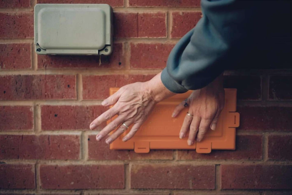 A person's hands manipulate an orange access panel on a red brick wall, with a second grey panel above it. Close-up, indoor.