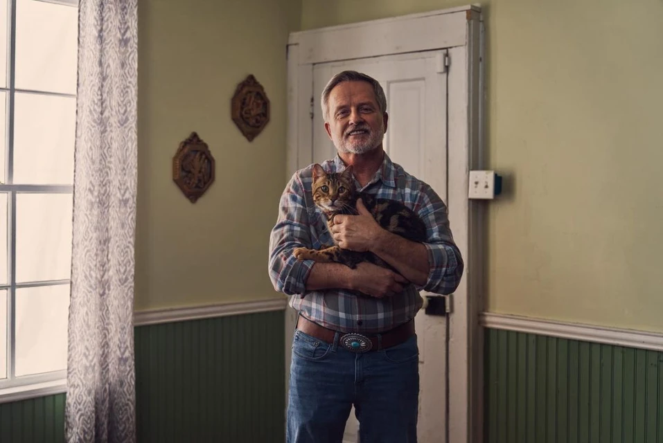 A middle-aged man with a gray beard is holding a tabby cat in an indoor setting, standing in front of a white door and green walls.
