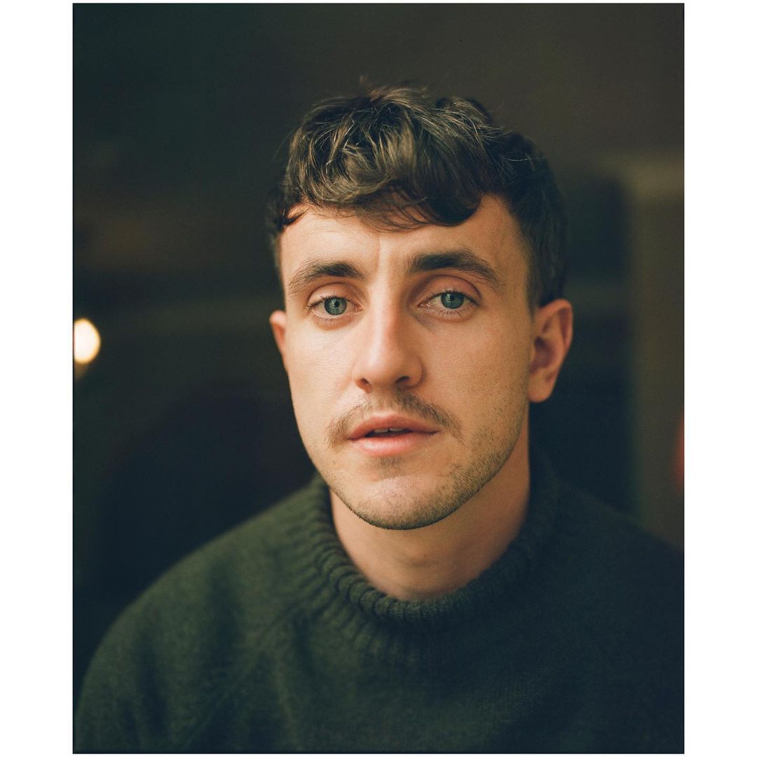 Close-up portrait of an adult male with dark hair and a moustache, wearing a green sweater against a blurred background.