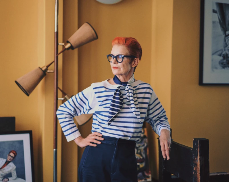 Stylish mature woman with orange hair, wearing a striped shirt and glasses, posing indoors with hands on hips, in front of a yellow wall.