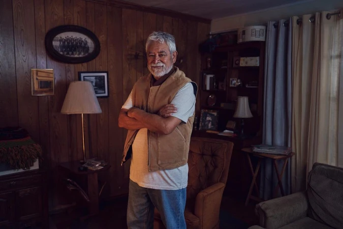 Portrait of an elderly Caucasian man indoors. He is wearing a vest, a t-shirt, and jeans, and has his arms crossed, smiling into the camera.