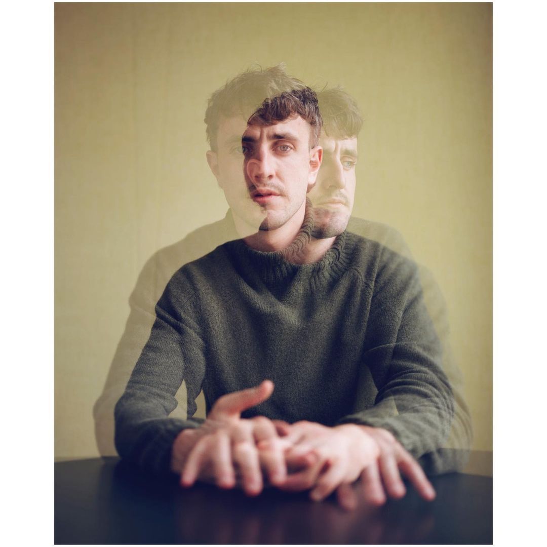Portrait of a man with a blurred, superimposed effect, wearing a green sweater and sitting at a dark-colored table against a pale yellow background.