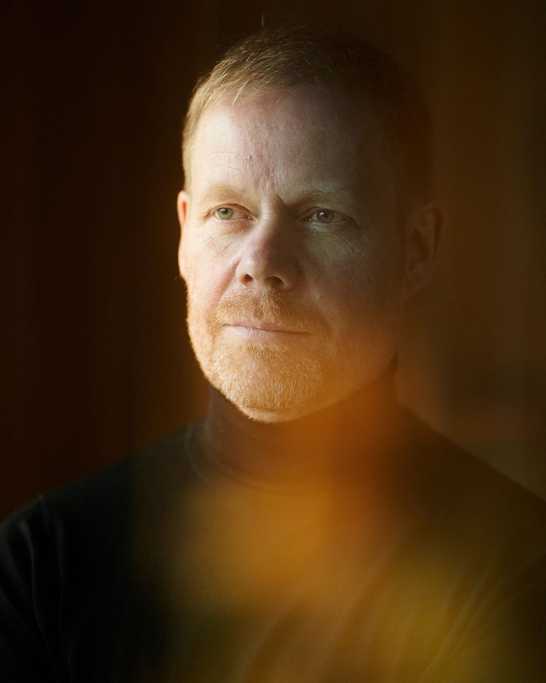 Portrait of a middle-aged man with fair skin and red hair, gazing slightly to the left against a dark background with soft lighting.