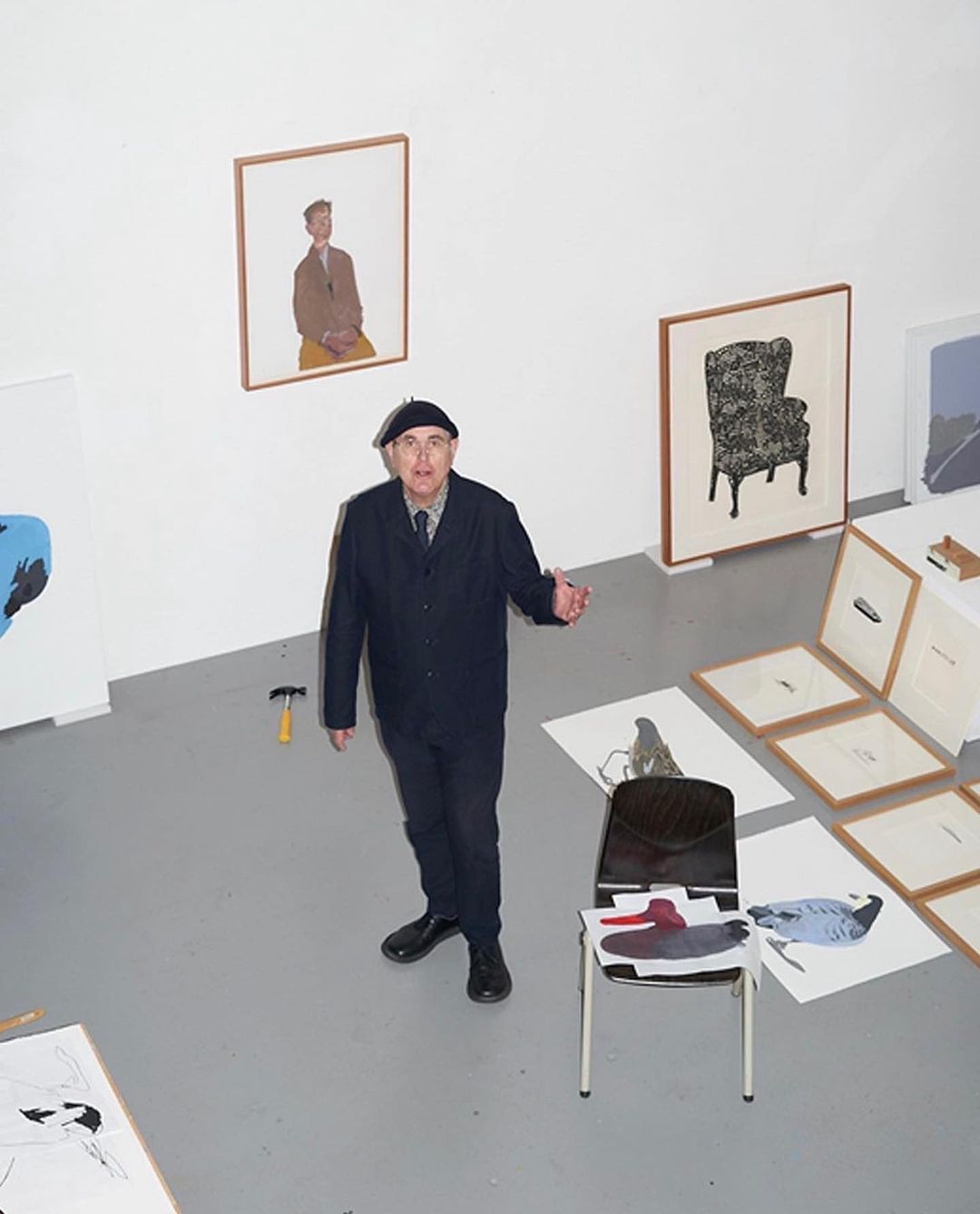 Overhead shot in art studio: senior man in beret stands among framed artworks, hammer, chair, and sketches; white walls and grey floor.