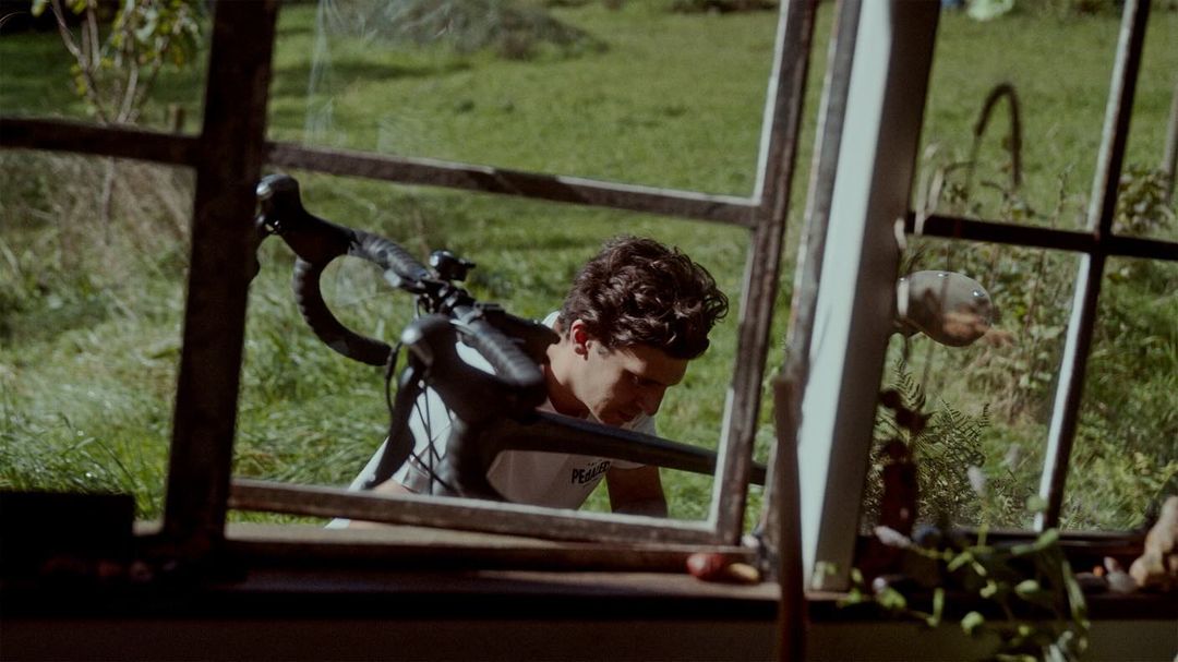 Man with curly hair and a white t-shirt looking down inside an old weathered window frame, with a bicycle resting on his shoulder.