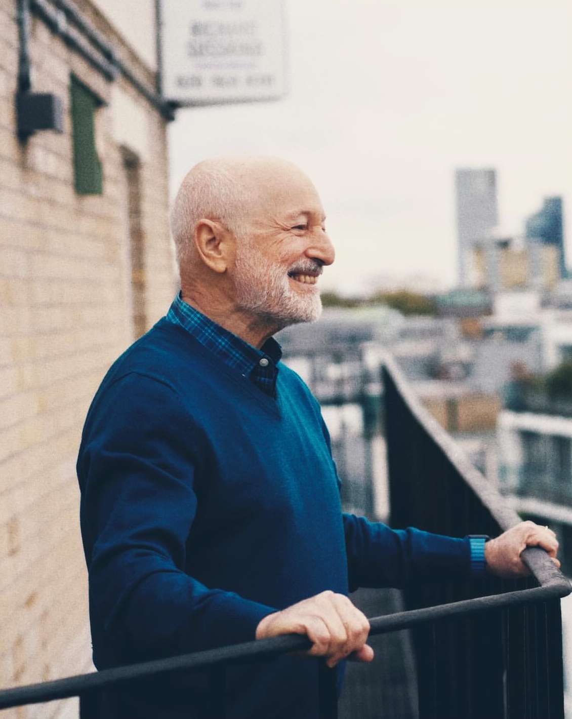 An elderly man with a beard stands on a balcony in a blue sweater, smiling, with a city visible in the background on a bright day.