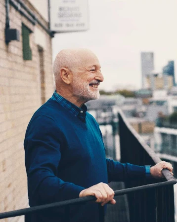 An elderly man with a beard stands on a balcony in a blue sweater, smiling, with a city visible in the background on a bright day.