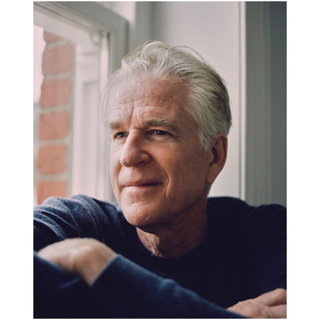 Portrait of a mature adult male with gray hair, wearing a dark sweater, looking towards a window with natural light indoors.