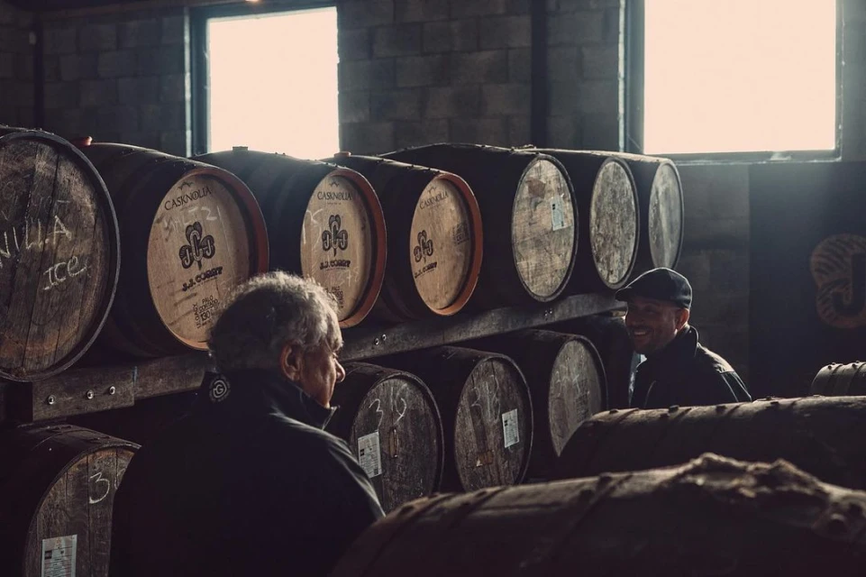 Two men in a dimly lit distillery cellar, standing among rows of aging whiskey barrels stacked high, creating a rustic ambiance.