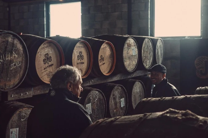 Two men in a dimly lit distillery cellar, standing among rows of aging whiskey barrels stacked high, creating a rustic ambiance.