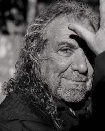 Close-up black and white portrait of an elderly man with long curly hair raising a hand to his forehead, photographed by Tom Jamieson.