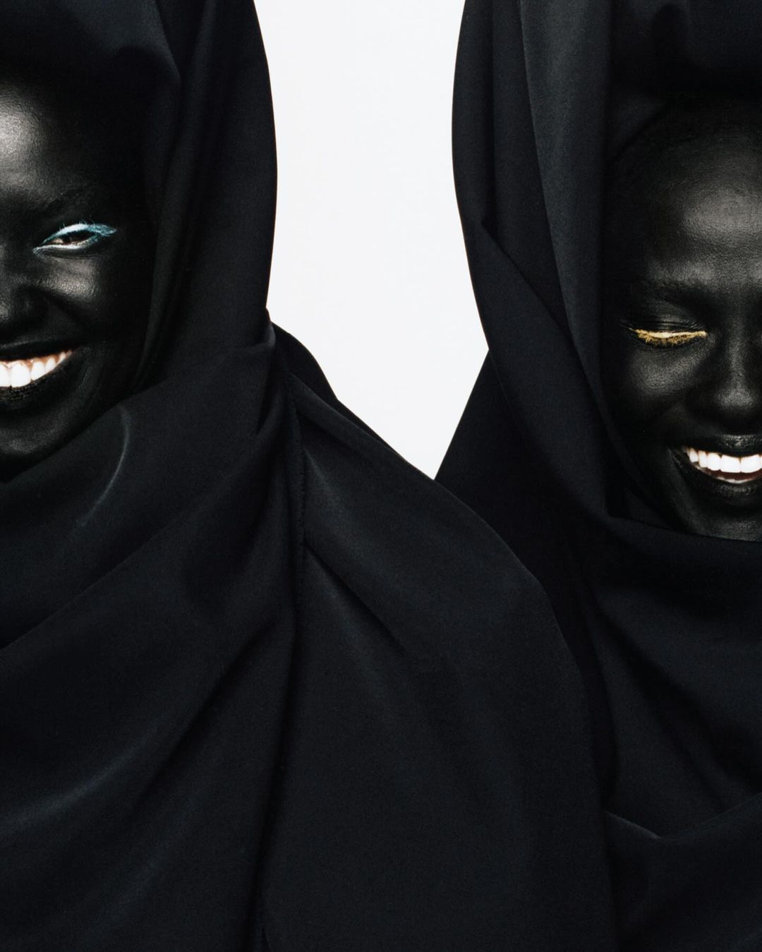 Two Black women smile, wearing black hoods, with blue and gold eye makeup against a white backdrop, creating a striking contrast and high fashion vibe.