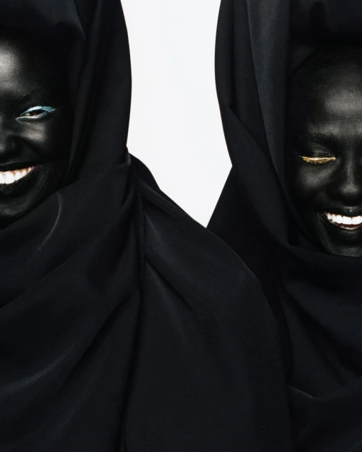 Two Black women smile, wearing black hoods, with blue and gold eye makeup against a white backdrop, creating a striking contrast and high fashion vibe.