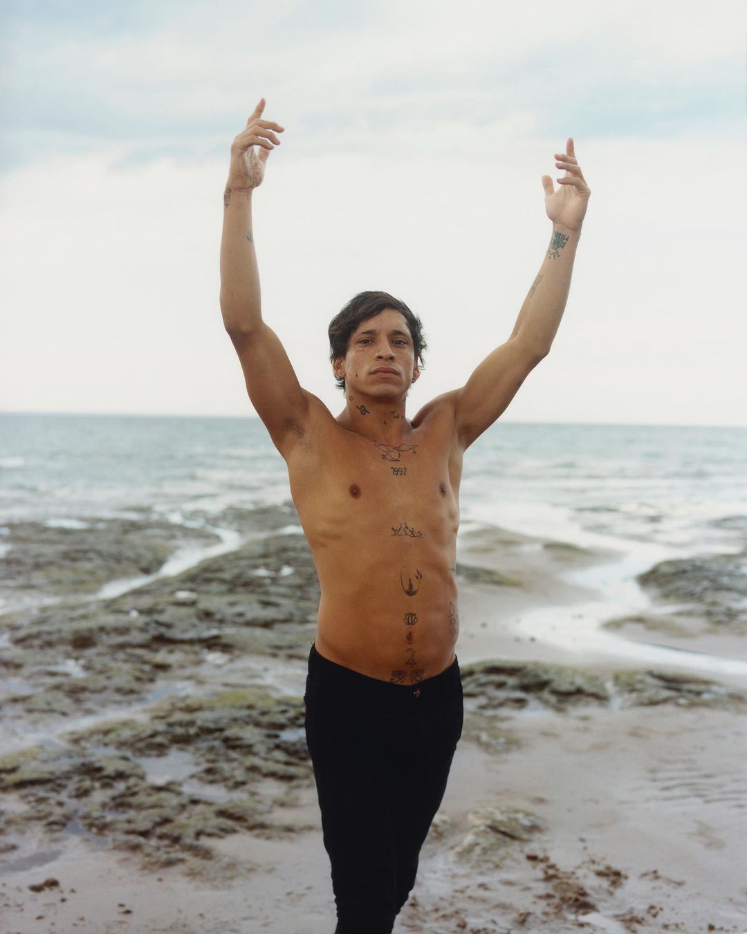 Man with tattoos standing shirtless on a beach with his arms raised against a cloudy sky and ocean in the background.