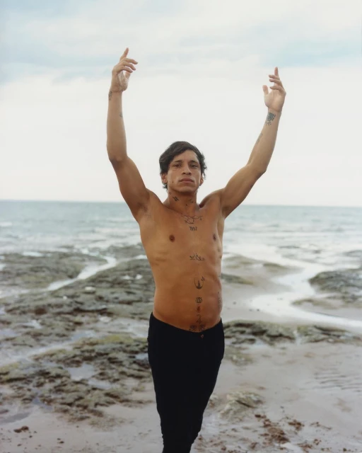 Man with tattoos standing shirtless on a beach with his arms raised against a cloudy sky and ocean in the background.